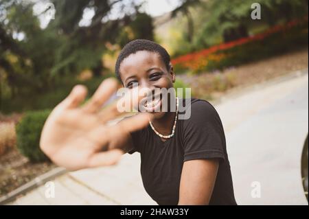 Primo piano ritratto di attraente felice sorridente giovane bellezza naturale capelli corti donna africana che indossa nero t-shirt ridere allungando le mani di chiusura dalla macchina fotografica nel parco naturale estivo Foto Stock