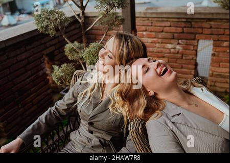 Primo piano ritratto di due amici femminili in stretto grigio abiti ridendo mentre si siede su sedie di vimini sulla terrazza all'aperto in strada caffè estate. Foto di alta qualità Foto Stock