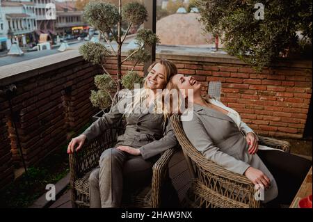 Primo piano ritratto di due amici femminili in stretto grigio abiti ridendo mentre si siede su sedie di vimini sulla terrazza all'aperto in strada caffè estate. Foto di alta qualità Foto Stock