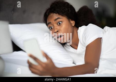 Scioccata donna nera guardando il cellulare, steso a letto Foto Stock