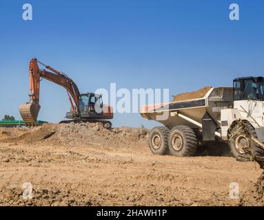 escavatore cingolato arancione e dumper articolato durante la fase di scavo in un cantiere. Carico e trasporto di suolo. Lavoro di co Foto Stock