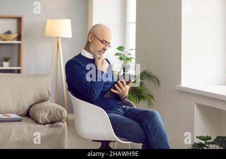 Uomo anziano seduto in una sedia a casa, tenendo un tablet, leggendo un ebook e pensando Foto Stock