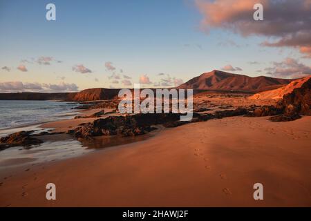 La costa occidentale di Lanzarote al tramonto. Una spiaggia con sabbia e rocce e un cielo blu con alcune nuvole. Vista sulla catena montuosa Los Ajaches. Isole Canarie Foto Stock