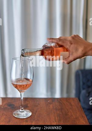 mano dell'uomo che versa il vino di rosa in un bicchiere su un tavolo di legno con una tenda bianca sullo sfondo a casa Foto Stock