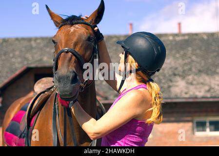 Donna rider che parla con il suo cavallo. Ritratto di cavallo con donna in casco. Cavallo equestre con pilota e vecchia stalla sullo sfondo. Foto Stock