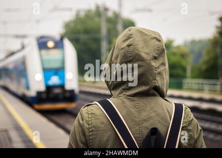 Turista irriconoscibile con cappa in attesa alla stazione ferroviaria al momento dell'arrivo del treno in pioggia. Trasporto ferroviario. Allontanarsi da tutto Foto Stock