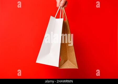 Mano femmina che tiene due borse shopping isolato su sfondo rosso. Sacchetti di carta bianca e marrone artigianali in mano. Offerta black friday, sconto, shopping Foto Stock