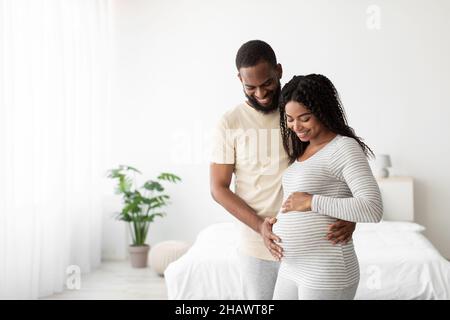 Sorridendo giovane africano americano marito abbraccia la moglie incinta, toccando il ventre in camera da letto minimalista interno Foto Stock