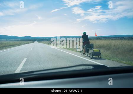 Biker sul percorso che tira il rimorchio da turismo della bicicletta. Strada di campagna con colline in fondo. Vista dall'interno della vettura Foto Stock