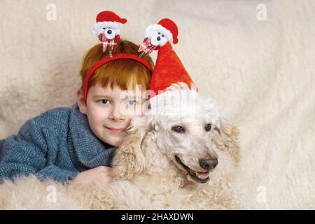 Un ragazzo giovane che indossa un cappello santa sorride e abbraccia un grande cane da cuccioli bianco reale. Il rapporto tra un cane e un bambino. Foto Stock