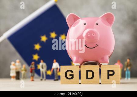 Piggy banca, cubi di legno con testo, giocattoli in plastica e una bandiera su sfondo astratto, un concetto sul tema del PIL dell'UE Foto Stock