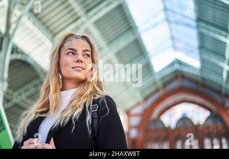 Basso angolo di giovane donna con capelli biondi ricci che indossano un cappotto nero e zaino in piedi in un edificio moderno Foto Stock