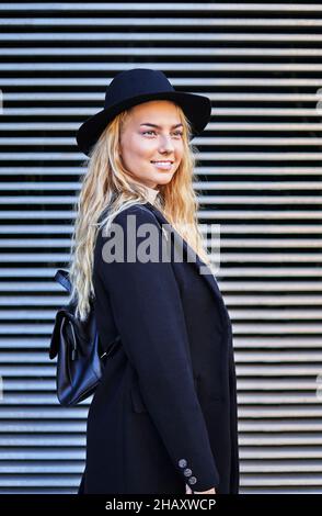 Modello femminile giovane bionda sorridente e alla moda con cappello nero e cappotto con zaino in pelle Foto Stock