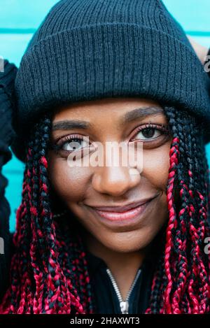 Giovane contenuto nero Donna con trecce Afro indossare bennie scuro e guardando la fotocamera su sfondo blu Foto Stock