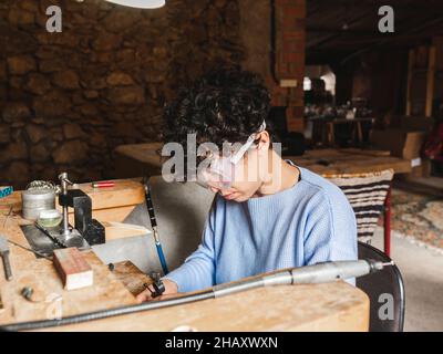 Da sopra vista laterale di lavoratrice etnica ricci pelata in vetri trasparenti che si concentrano al banco da lavoro orafo Foto Stock