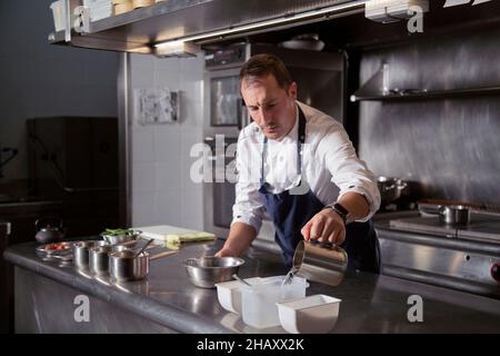 Chef professionista concentrato con tazza in acciaio versando acqua nel contenitore mentre si preparano gli ingredienti per il processo di cottura in cucina del ristorante Foto Stock