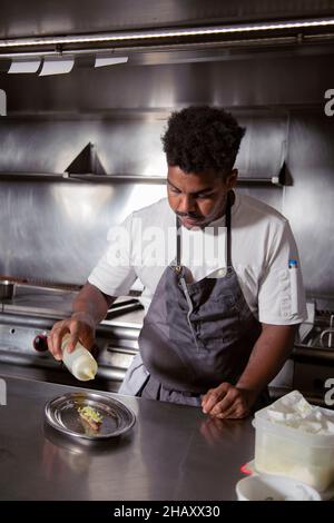 Cuoco maschile africano concentrato americano in grembiule versare olio al piatto di alta cucina con pesce in cucina del ristorante Foto Stock