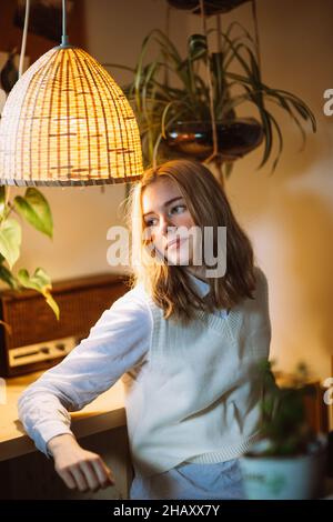 Giovane donna in blusa bianca seduta alla scrivania sotto la lampada a sospensione incandescente in camera con fiori in vaso Foto Stock