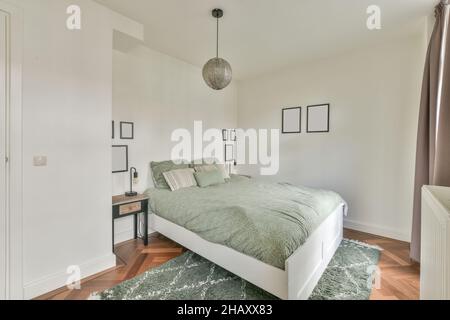 Confortevole camera da letto con moquette e letto coperto con copriletto e cuscini comodini quadri su pareti bianche e lampada a sospensione Foto Stock