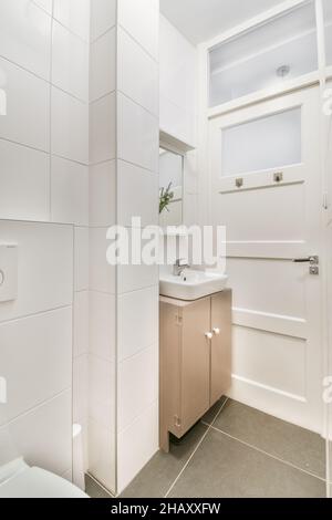 Interno di uno stretto bagno bianco con ciotola bianca e lavandino in ceramica con specchio collocato vicino a una parete piastrellata Foto Stock