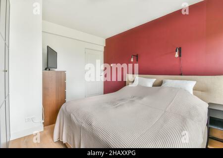 Comodo letto con coperta posto vicino a TV moderna su cassetti in camera da letto luminosa con armadio grigio costruito vicino alle finestre in appartamento Foto Stock