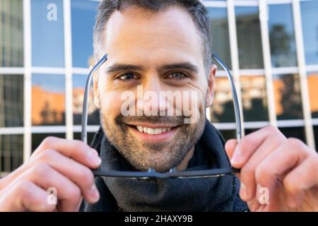 Coltivare allegro maschio in abiti casual sorridendo e indossare occhiali sulla strada contro edificio moderno di giorno Foto Stock