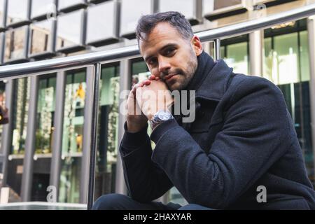 Uomo in cappotto nero seduto sulle scale vicino ringhiera di metallo sulla strada e guardando la macchina fotografica contro l'edificio contemporaneo di giorno Foto Stock