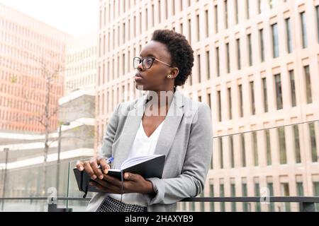 Un'esperta femminile afro-americana che prende appunti in notebook mentre si alza sul balcone di un moderno edificio per uffici Foto Stock
