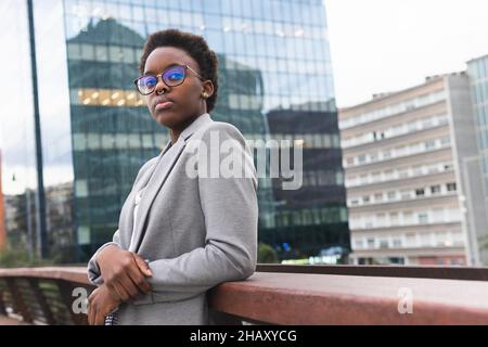 Giovane manager donna nera che indossa abiti alla moda e occhiali appoggiati sulla ringhiera mentre riposa nel balcone dell'edificio moderno Foto Stock