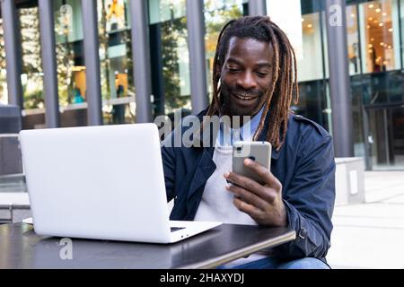 Messaggi positivi di giovane uomo afroamericano su smartphone seduti a tavola con un computer portatile sulla terrazza del centro commerciale Foto Stock
