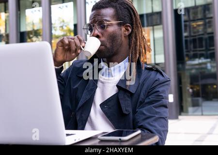 Serio imprenditore africano americano maschile in occhiali e con i doadlock in abiti casual bere caffè mentre si lavora su laptop sittin remoto Foto Stock