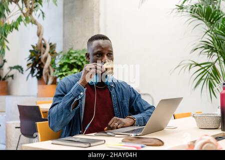 Serio lavoratore di sesso maschile afroamericano con cuffie che bevono bevande calde mentre si siede alla scrivania con netbook moderno durante il lavoro in ufficio Foto Stock