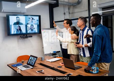 I colleghi multietnici agitano le mani mentre si terrà una videoconferenza in una sala riunioni leggera con tavolo e computer portatili durante le riunioni in un ufficio moderno Foto Stock