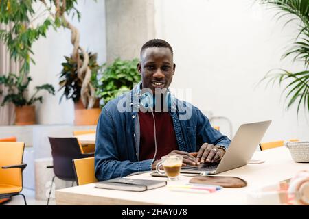 Concentrò il lavoratore maschile afroamericano con le cuffie durante la digitazione sul netbook al desktop con il caffè durante il lavoro in ufficio guardando la fotocamera Foto Stock