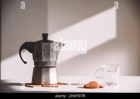 Pentola di moka in metallo posta su un tavolo bianco vicino a una tazza di vetro e biscotti a forma di cuore nelle giornate di sole Foto Stock
