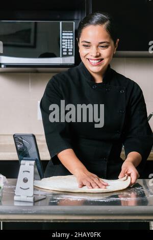 Giovane e sorridente chef etnico femminile in uniforme che prende video sullo smartphone mentre cucinano deliziosa pizza sul banco della cucina Foto Stock