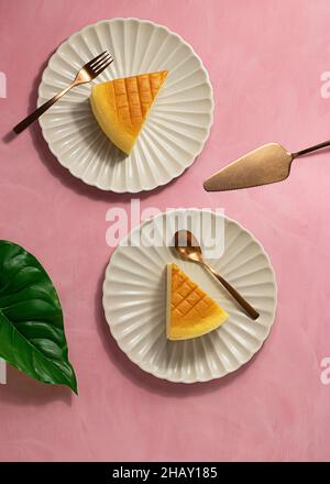 Vista dall'alto dei piatti con fette di gustosa cheesecake giapponese e argenteria poste vicino alla spatola e foglia verde di piante su sfondo rosa Foto Stock