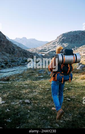 Vista posteriore del viaggiatore irriconoscibile con zaino che cammina su terreni erbosi con sentieri e pietre durante il viaggio nella natura Foto Stock