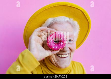 Donna anziana felice con capelli grigi in elegante cappello giallo coprente occhio con ciambelle dolci glassate rotonde su sfondo rosa Foto Stock