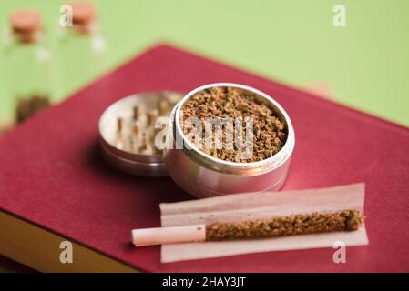 marijuana tritata su macinacaffè, su carta di sigaretta con filtro e bottiglia di vetro ermetica, processo per la produzione di sigari marijuana. Sfondo rosso Foto Stock