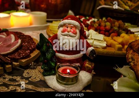 La statuina di Babbo Natale con un albero di Natale verde e una candela che brucia su un tavolo festivo con una varietà di prelibatezze di carne e formaggio su vassoi di legno Foto Stock