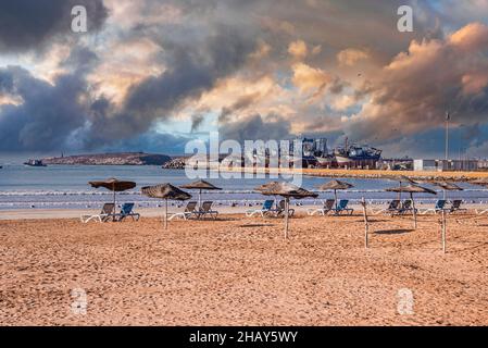 Sedie da spiaggia con ombrelloni in fila sulla spiaggia sabbiosa in giornata di sole contro il cielo nuvoloso Foto Stock