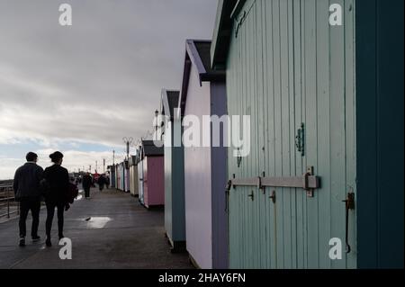 Felixstowe, Suffolk, UK : la gente cammina lungo il lungomare di Felixstowe in un inverno freddo e noioso passato una linea di capanne di spiaggia di fronte al mare. Foto Stock