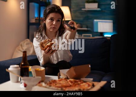 Giovane donna seduta sul divano mangiare doppio cheeseburger tenendo telecomando girare volume in televisione guardare le notizie della sera. Persona che cambia i canali tv mentre mangia hamburger da asporto. Foto Stock