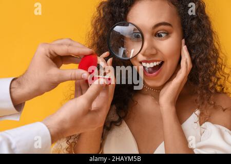 Felice giovane donna afroamericana che guarda l'anello di fidanzamento dato dal suo ragazzo attraverso la lente di ingrandimento su sfondo a colori Foto Stock