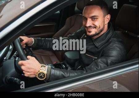 Ritratto di bel giovane uomo sorridente mentre guida auto moderna. Autista che indossa abiti casual e orologio dorato. Foto Stock