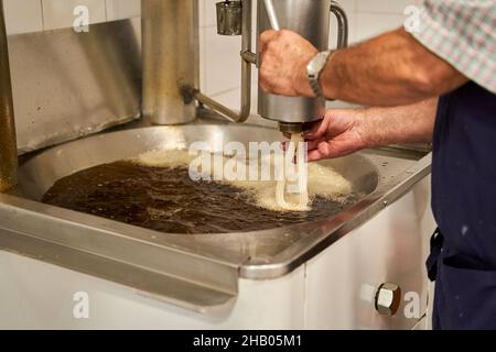 Vista laterale di un cuoco non identificato in grembiule versare l'impasto crudo in olio fritto mentre si preparano churros, tipica colazione spagnola. Foto Stock