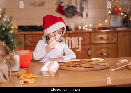 Home panetteria, cucina tradizionale dolci festive. Bambina in rosso santa Hat preparazione per la preparazione di pasta di pan di zenzero su tavola di legno. Anno nuovo Foto Stock