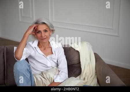 Sorridente donna matura ed elegante con capelli grigi seduti sul divano e appoggiati a portata di mano mentre si rilassa in salotto e si guarda la macchina fotografica Foto Stock