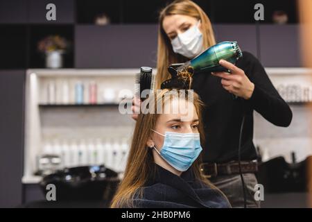 Master donna parrucchiere in una maschera medica asciuga i capelli della ragazza con un asciugacapelli e pettini dopo il lavaggio in un salone di bellezza. Covid-19 pandemia e. Foto Stock